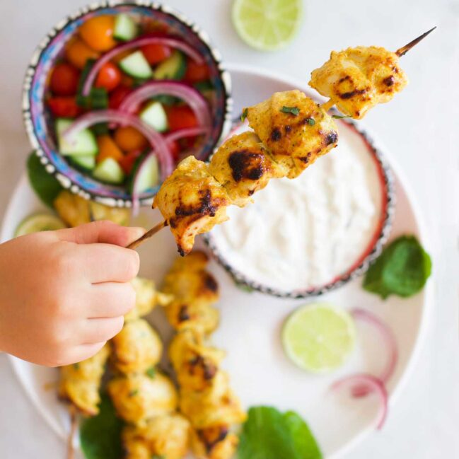 Child Holding a Chicken Tikka Skewer with Pate of Skewers, Dip and Salad in Background