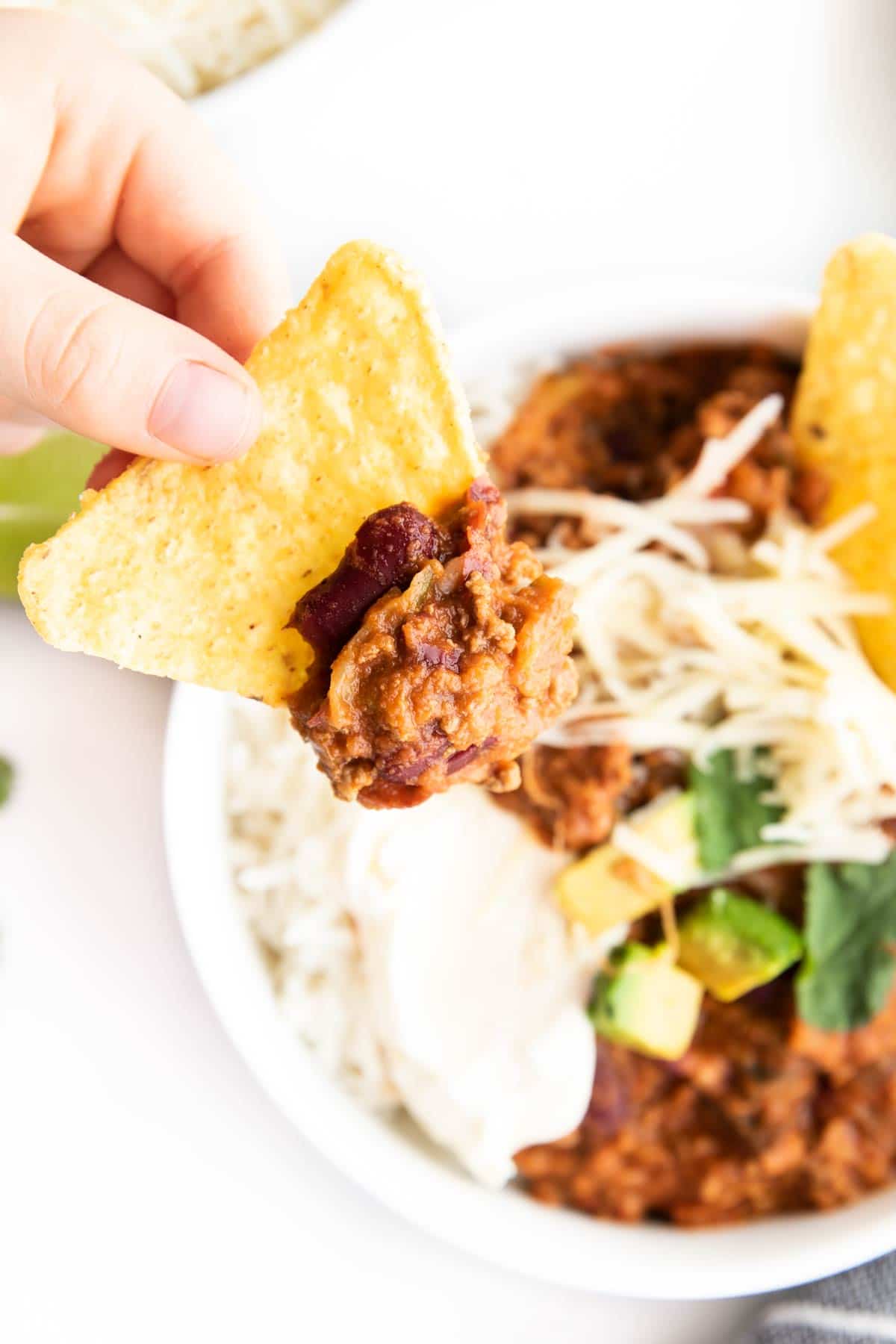 Child's Hand Holding Tortilla Chip Topped with Chilli With Bowl of Chilli in Background