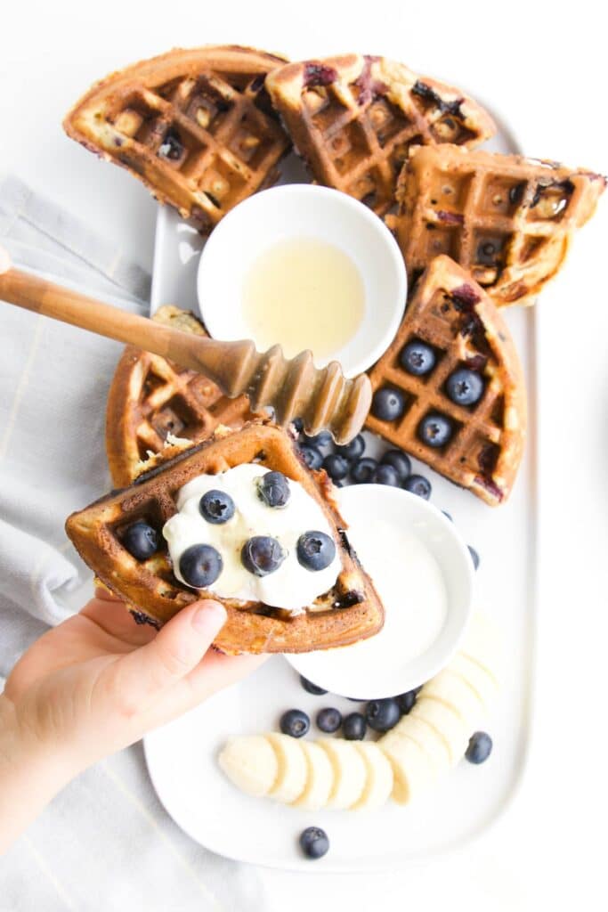 Child's Hand Holding Waffle with Yogurt and Blueberry Topping. Plate of Waffles and toppings in background.