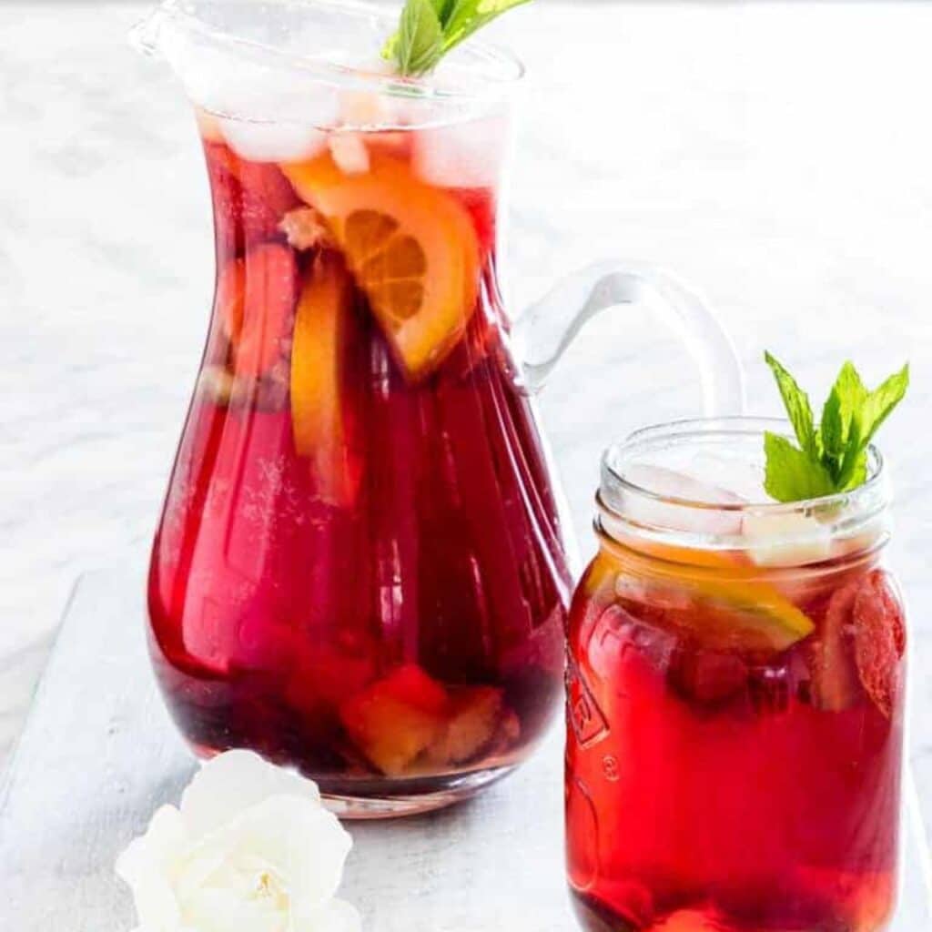 Jug of Sangria Mocktail with Fresh Fruit in Background with Jar of the Mocktail in Foreground.