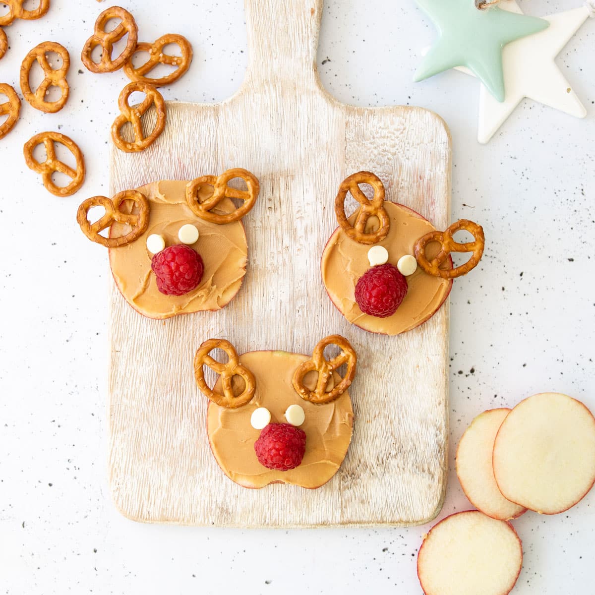 Three Slices of Apple Spread with Peanut Butter and Then Topped With Pretzels as Antlers, a Raspberry Nose and White Chocolate Drop Eyes. 