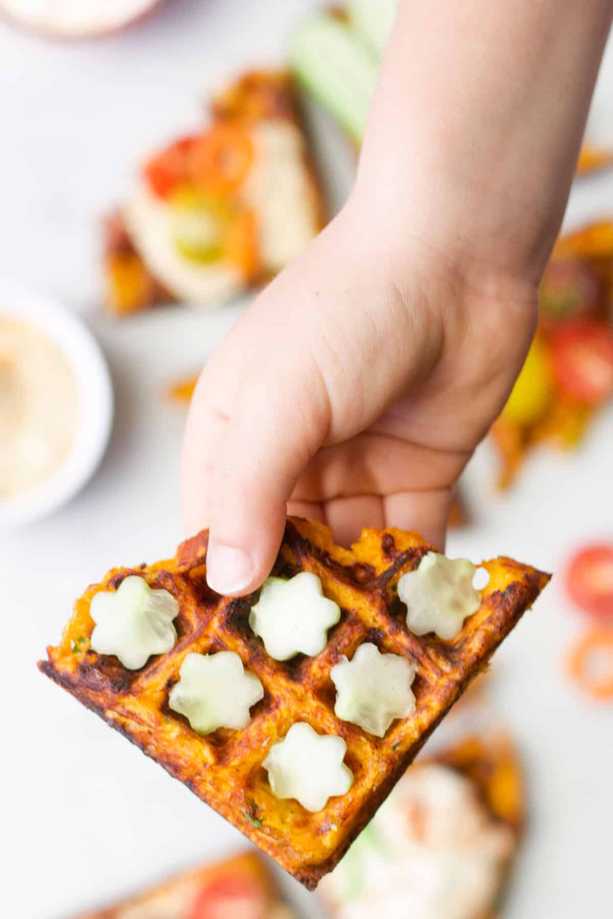 Child's hand holding savoury waffle topped with cucumber flowers. 