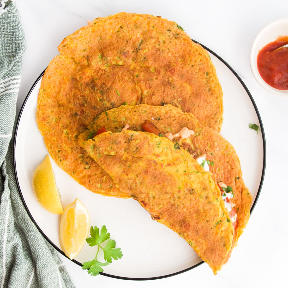 White Plate With Two Large Chickpea Pancakes On. One Folded Over. Lemon  on the Plate and Green Cloth in Background. 