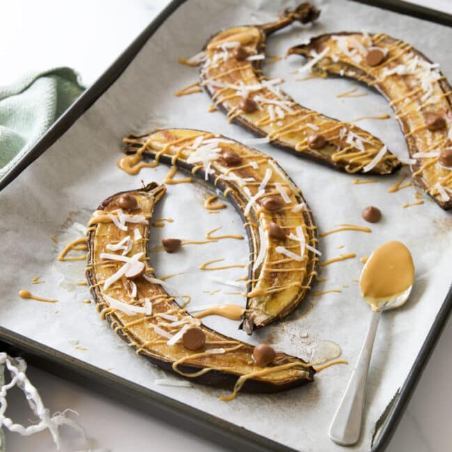 Baked Bananas on Baking Tray Topped With Peanut Butter Drizzle, Coconut Flakes and Choc Chips.