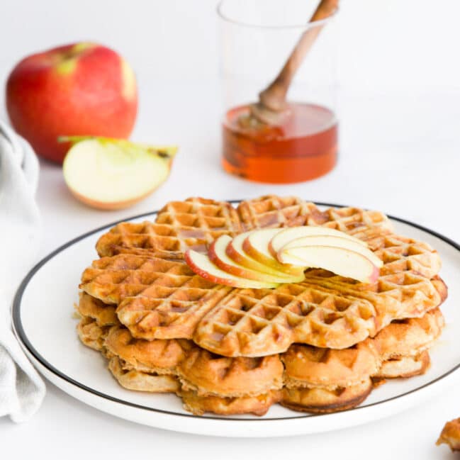 Stack of Apple Waffles on Plate Topped with Sliced Apple. Apple and Syrup in Background.