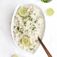 Coconut Lime Rice in White Serving Bowl with Squeezed Lime Halves and Sprinkled with Cilantro.