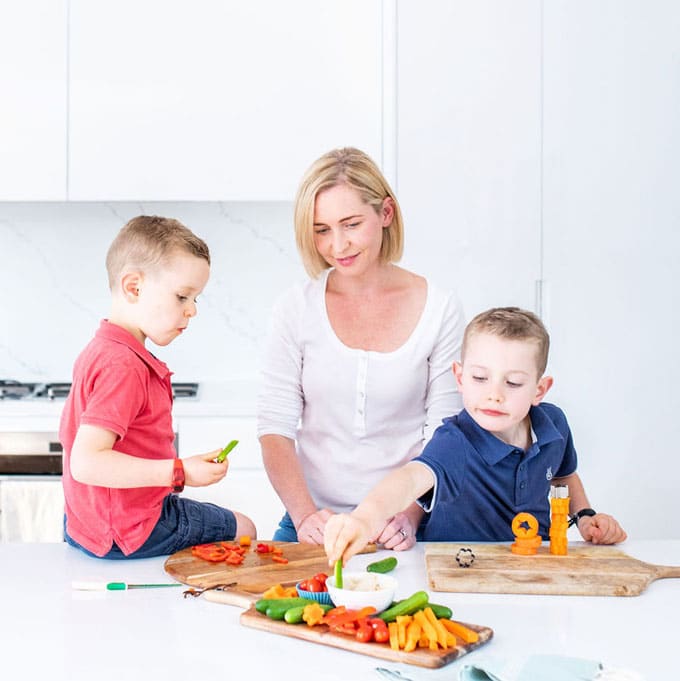 Amy in the kitchen with her two sons preparing and eating finger vegetables on wooden trays.