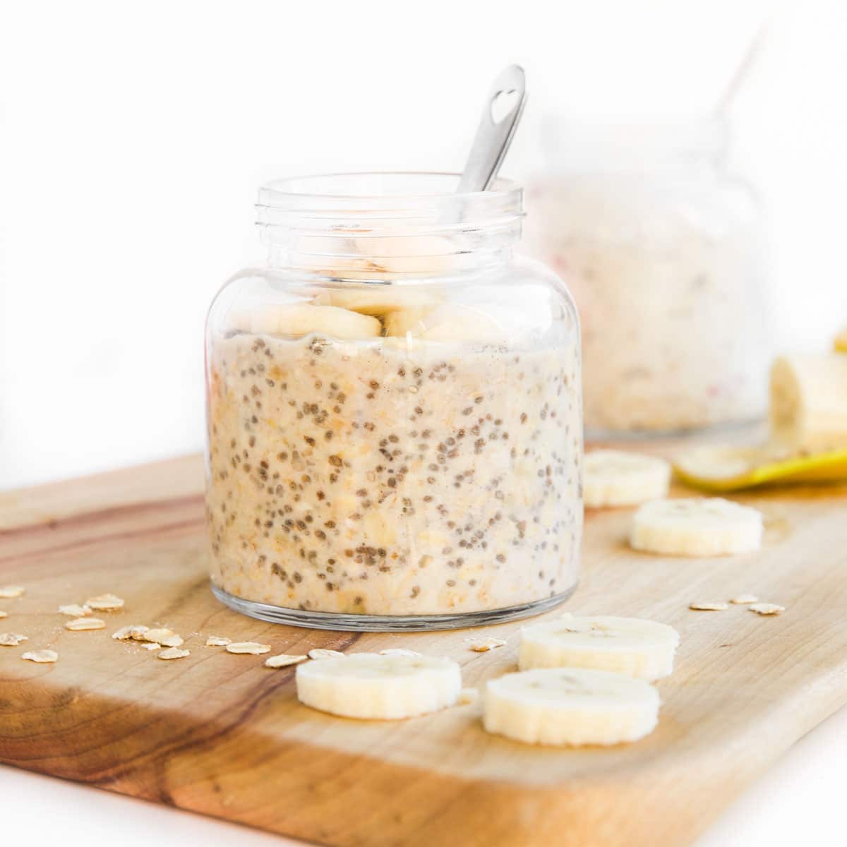 Banana Overnight Oats in Glass Jar Sitting on Wooding Chopping Board with Slices of Banana and Oats Scattered.