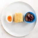 Top Down View of Plate with Half an Egg, Mini Corn on Cob and Silicon Muffin Case Filled with Grapes