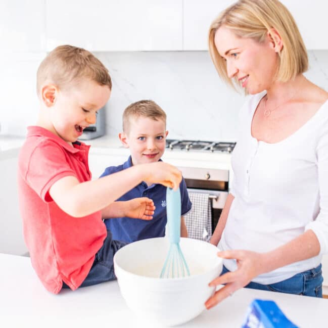 Mum in Kitchen with Two Boys. One boy sitting on Counter Top Stirring Mixture in Bowl