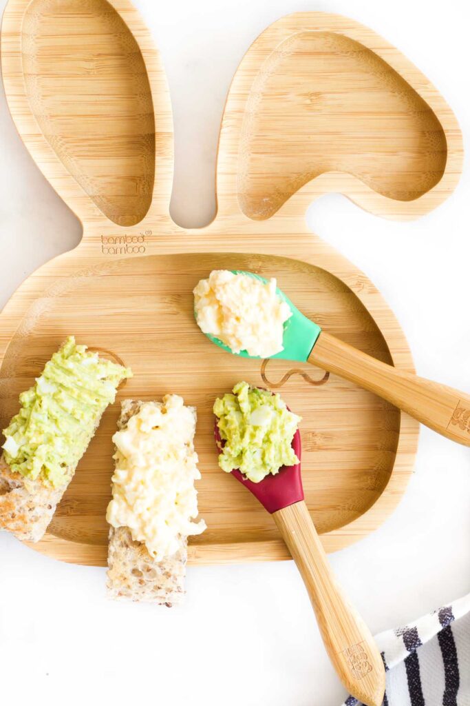 Baby Plate with Mashed Egg and Avocado on a Spoon and Spread on a Toast Finger.