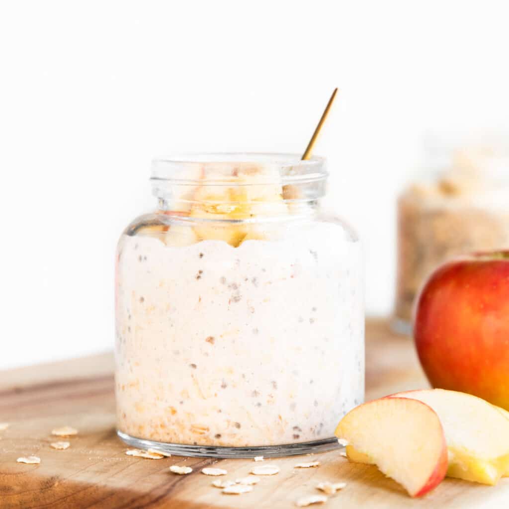 Jar of Apple Overnight Oats on Wooden Board with and Apple and Slices of Apple Scattered Next to it.