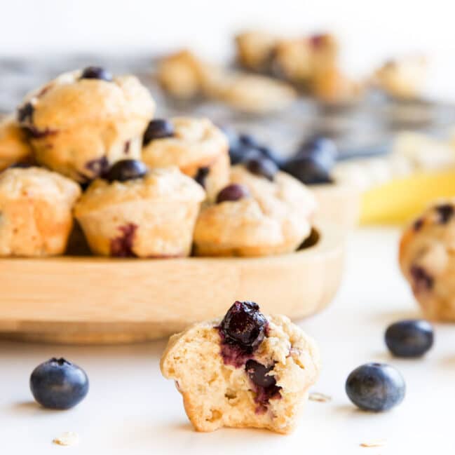 Side on Shot of Mini Blueberry Muffin with Bite Removed. A Plate of Muffing in Background and Muffin Tray