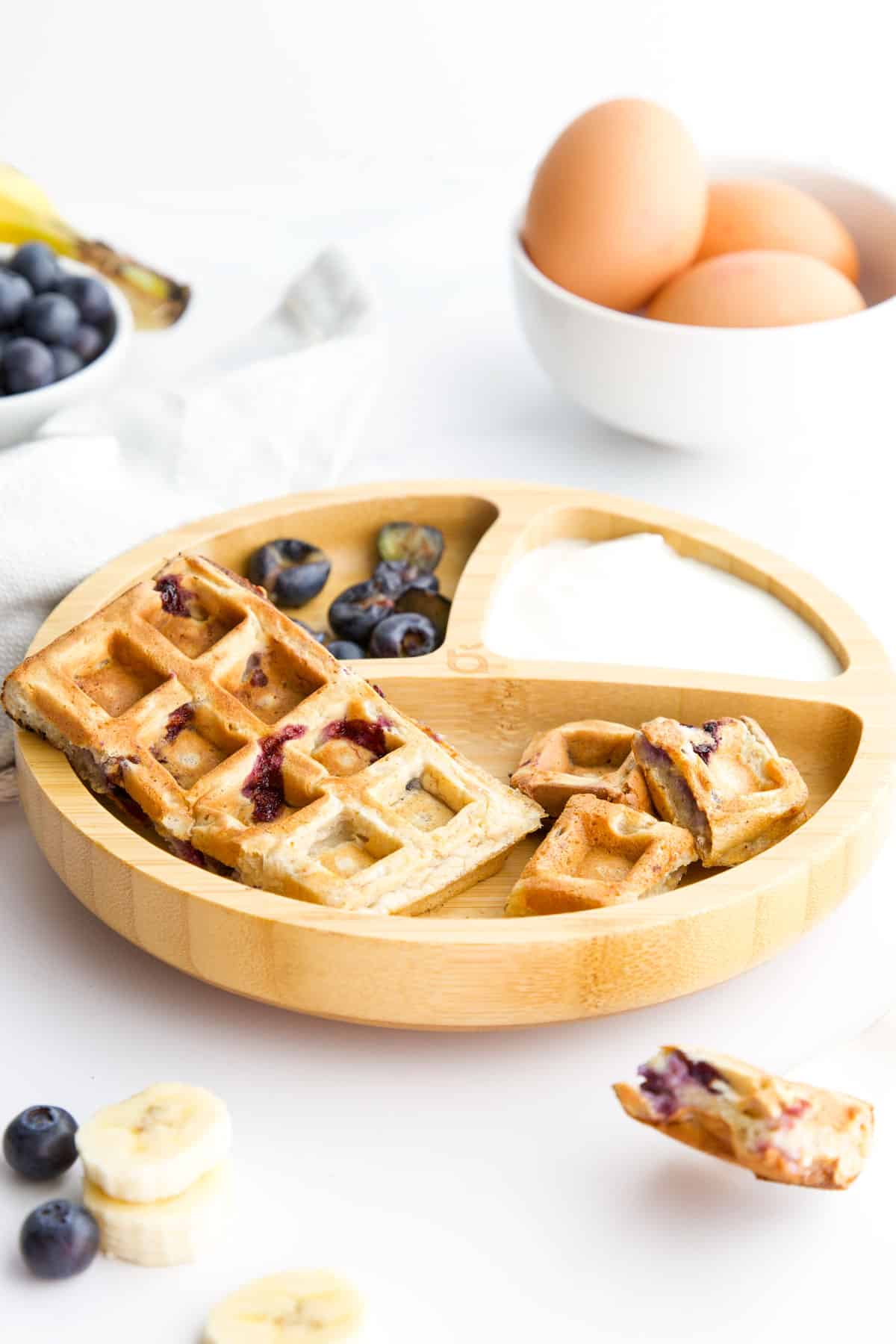 Baby Plate with Waffle Fingers and Waffle Squares Served with Blueberries and Yogurt. Eggs, Banana and Blueberries in the Background.
