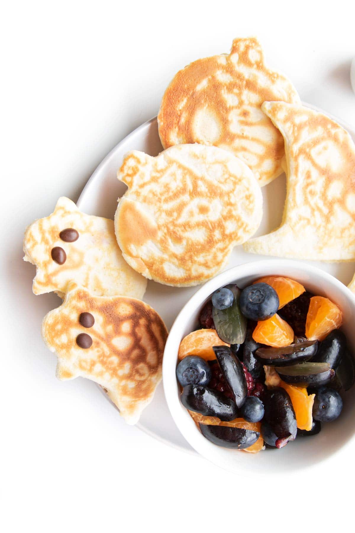 Plate of Halloween Shaped Pancakes (Ghost, Pumpkin, Hat) and Side of Fruit Salad