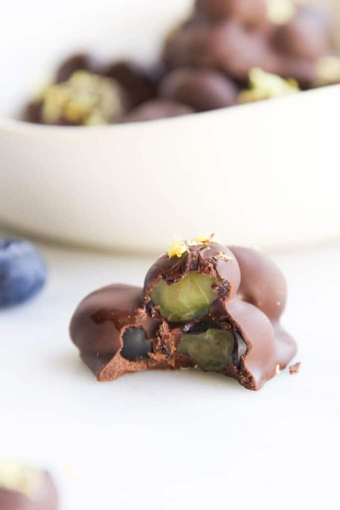 Side Shot of Chocolate Blueberry Cluster with Bite Taken Out. Plate of Chocolate Blueberries in Background