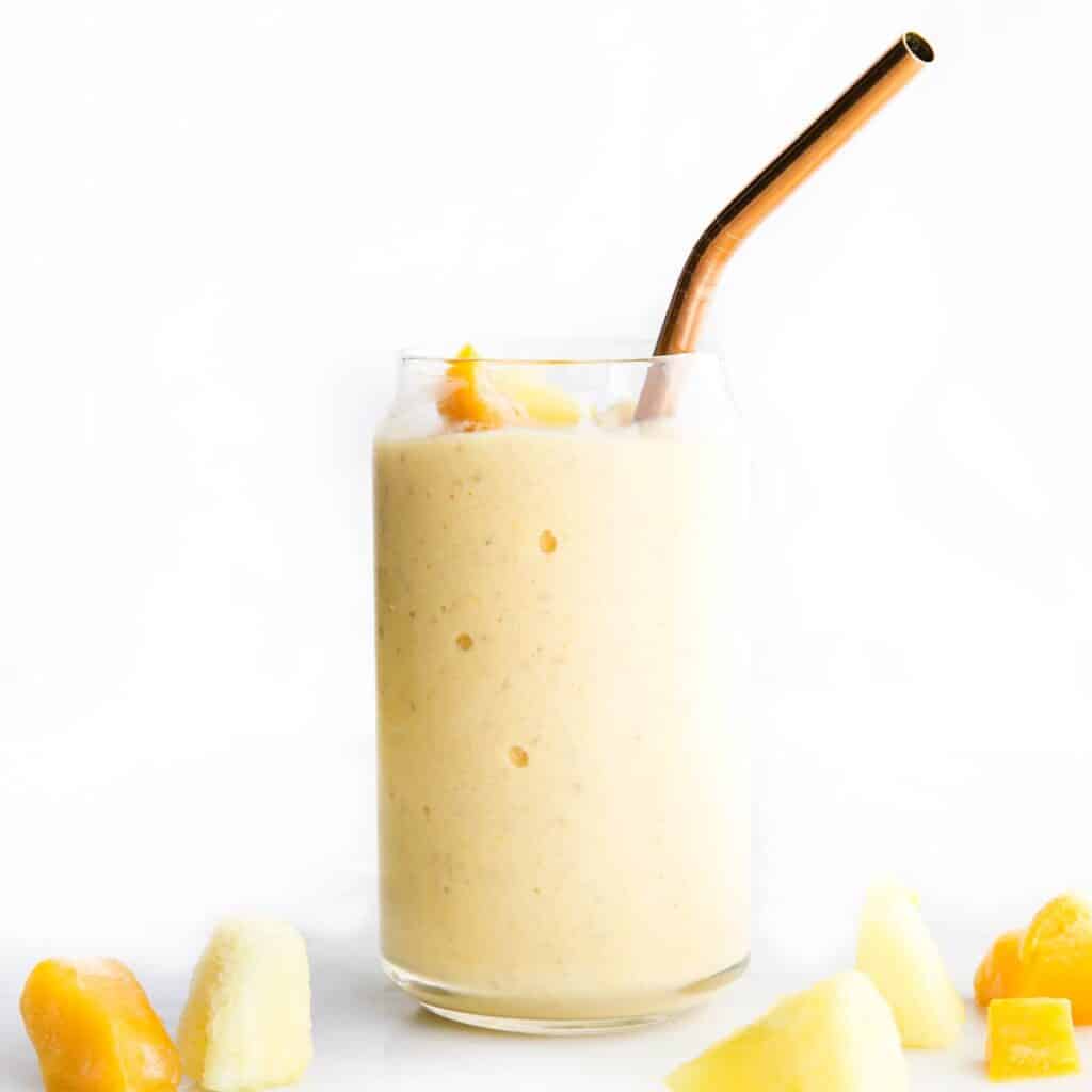 Side Shot of a Glass of Mango Pineapple Smoothie with Broze Straw Pieces of Mango anf Pineapple Scattered in Forefront