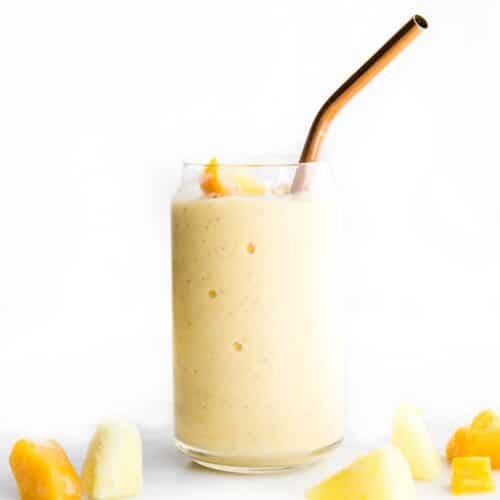 Side Shot of a Glass of Mango Pineapple Smoothie with Broze Straw Pieces of Mango anf Pineapple Scattered in Forefront