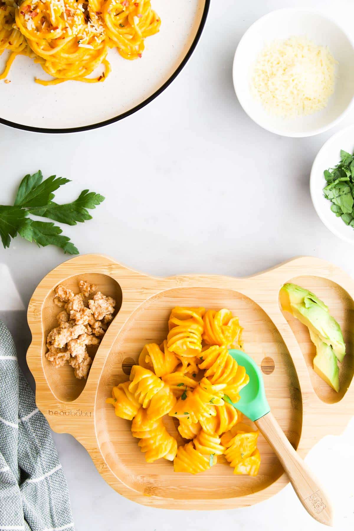Bamboo Koala Baby Plate with Carrot Pasta, Avocado and Chicken. Adult Plate and Bowls of Parmesan and Parsley in Background.