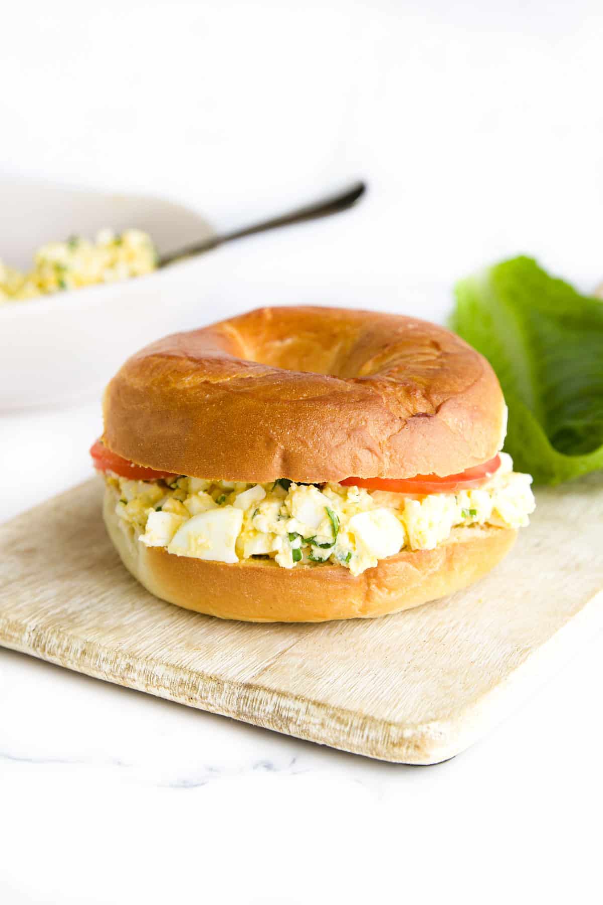 Cottage Cheese and Egg Salad Bagel Sitting on Wooden Board with Bowl of Cottage Cheese Egg Salad in Background.